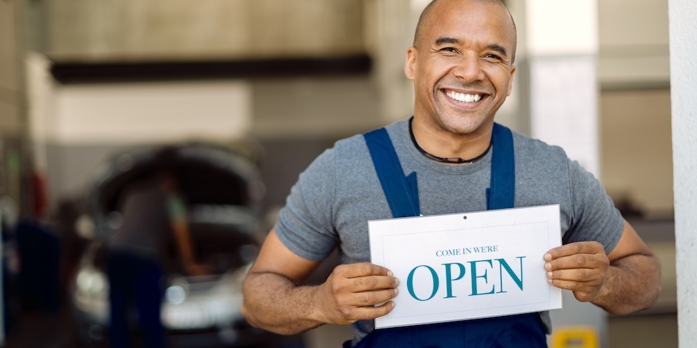 An auto repair shop technician holding a sign that says 'Come In! We're Open.'